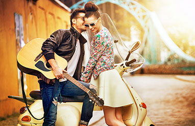 Junges Paar auf einer sonnigen Straße. Mann mit Gitarre auf Vespa. Frau mit Sonnenbrille und sommerlichem Outfit.